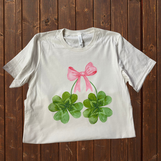 A vintage white unisex jersey t-shirt with a pink bow and green shamrock graphics on the front.