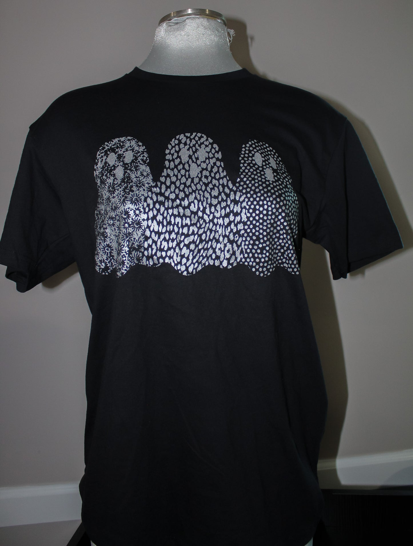 A black short-sleeve t-shirt displayed on a mannequin torso, featuring a large white graphic of three ghosts made of dense dots and lines across the chest.