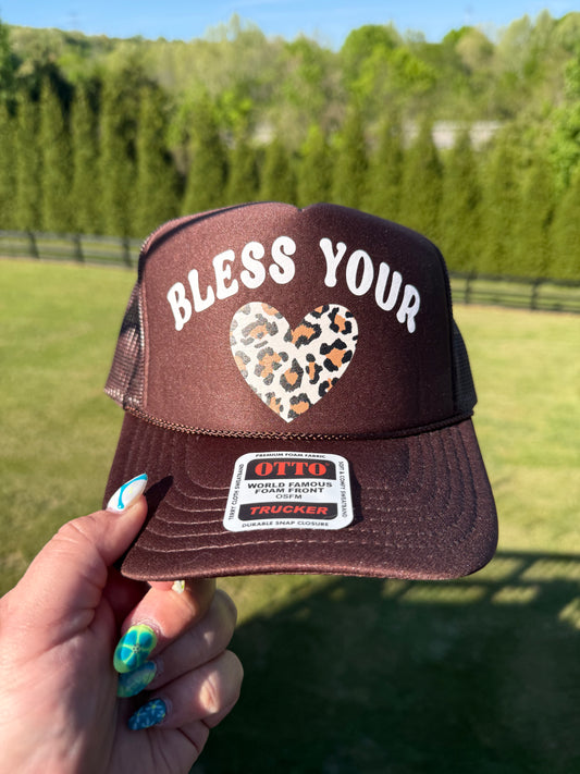 Brown trucker hat with 'Bless Your' and leopard print heart design held by a hand outdoors.