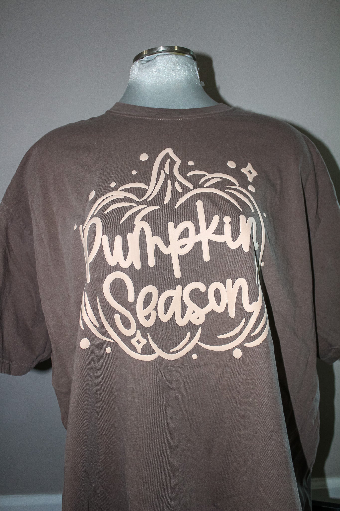 A brown short-sleeve t-shirt displayed on a mannequin torso, featuring a large cream-colored graphic on the front that reads 'Pumpkin Season' inside a pumpkin outline.