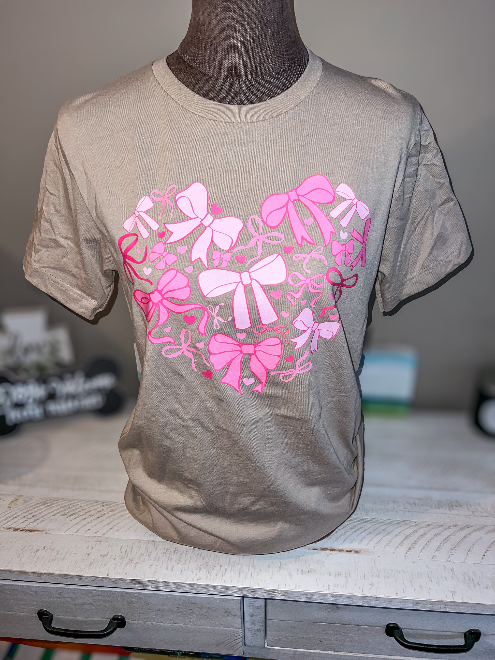 A beige T-shirt with a graphic of pink bows arranged in a heart shape on the front.