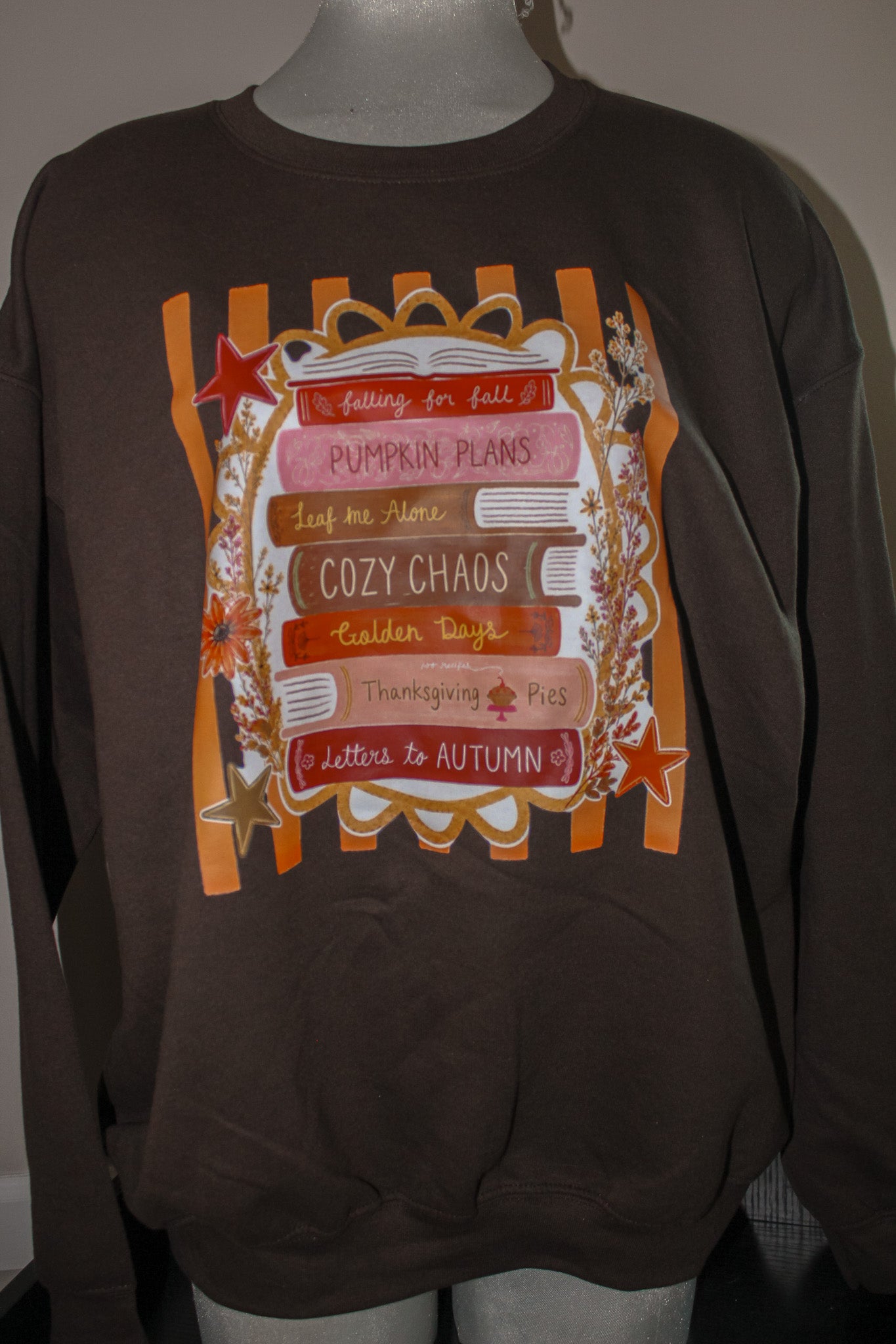 A dark brown crewneck sweatshirt displayed on a mannequin torso, featuring a large autumn-themed graphic print with orange stripes, stars, and layered pink and brown labels with fall phrases.