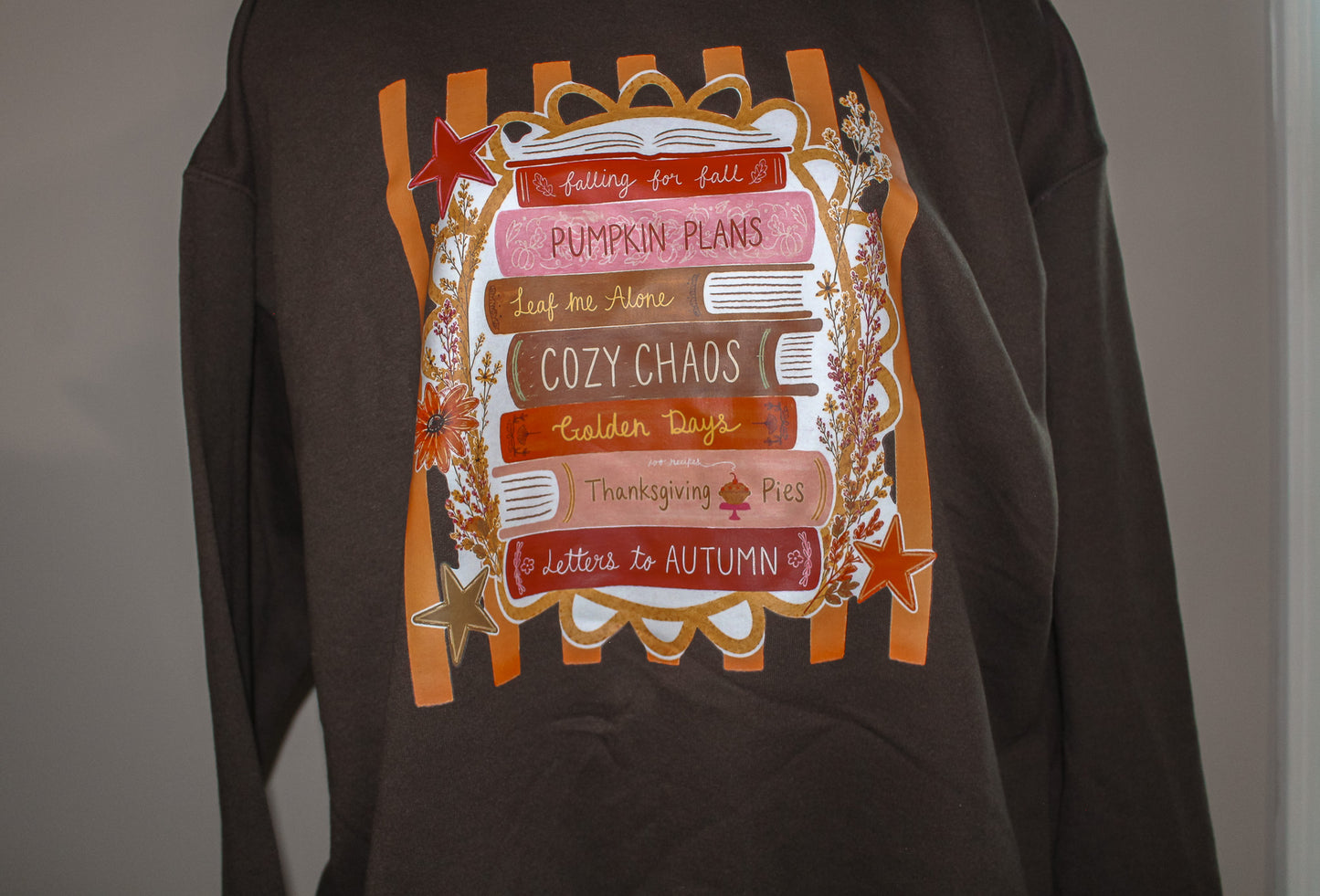 A dark brown long-sleeve sweatshirt featuring a large autumn-themed graphic print on the front with orange stripes, stacked books, stars, and phrases like 'Pumpkin Plans' and 'Cozy Chaos'.