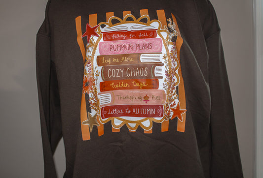 A dark brown long-sleeve sweatshirt featuring a large autumn-themed graphic print on the front with orange stripes, stacked books, stars, and phrases like 'Pumpkin Plans' and 'Cozy Chaos'.