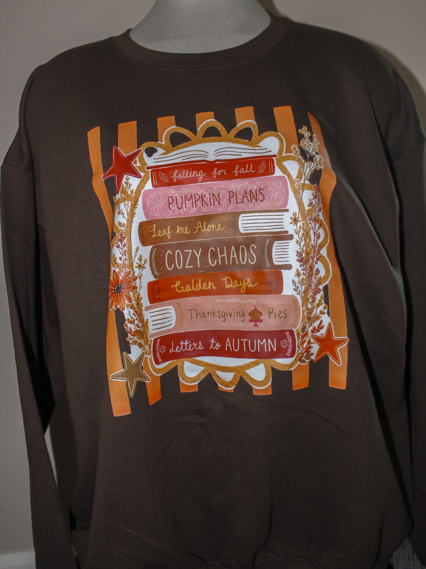 A dark brown long-sleeve t-shirt displayed on a mannequin torso, featuring a large autumn-themed graphic print with orange stripes, a book, stars, and text including 'Pumpkin Plans', 'Cozy Chaos', and 'Thanksgiving Pie'.