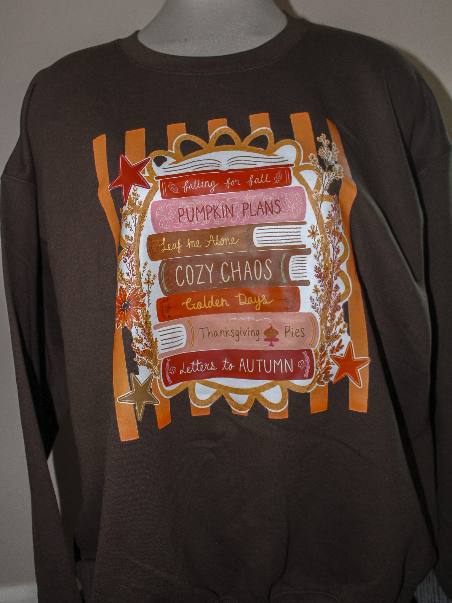 A dark brown long-sleeve t-shirt displayed on a mannequin torso, featuring a large autumn-themed graphic print with orange stripes, a book, stars, and text including 'Pumpkin Plans', 'Cozy Chaos', and 'Thanksgiving Pie'.