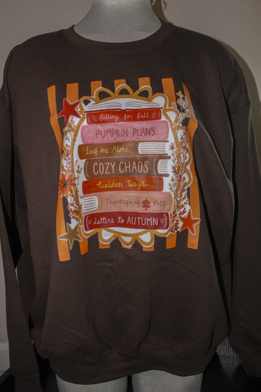 A dark brown crewneck sweatshirt displayed on a mannequin torso, featuring a large autumn-themed graphic print with orange stripes, stars, and layered pink and brown labels with fall phrases.