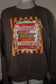 A dark brown crewneck sweatshirt displayed on a mannequin torso, featuring a large autumn-themed graphic print with orange stripes, stars, and layered pink and brown labels with fall phrases.