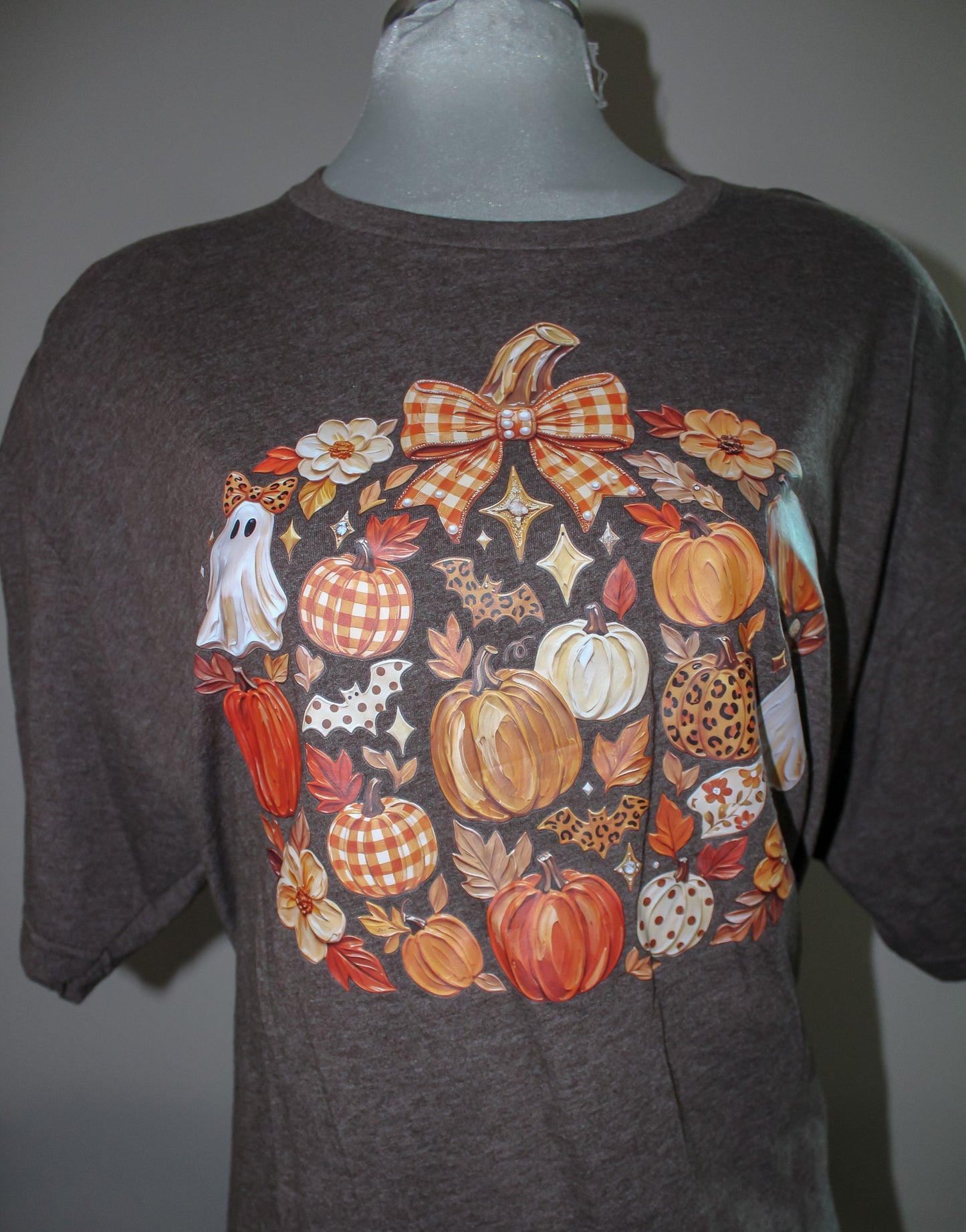 A dark brown heather t-shirt displayed on a gray mannequin torso, featuring a large autumn-themed graphic print with pumpkins, gingham patterns, a ghost, and leaves.
