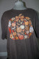 A dark brown heather short-sleeve t-shirt displayed on a mannequin torso, featuring a large autumn-themed graphic print with pumpkins, ghosts, flowers, and gingham patterns.