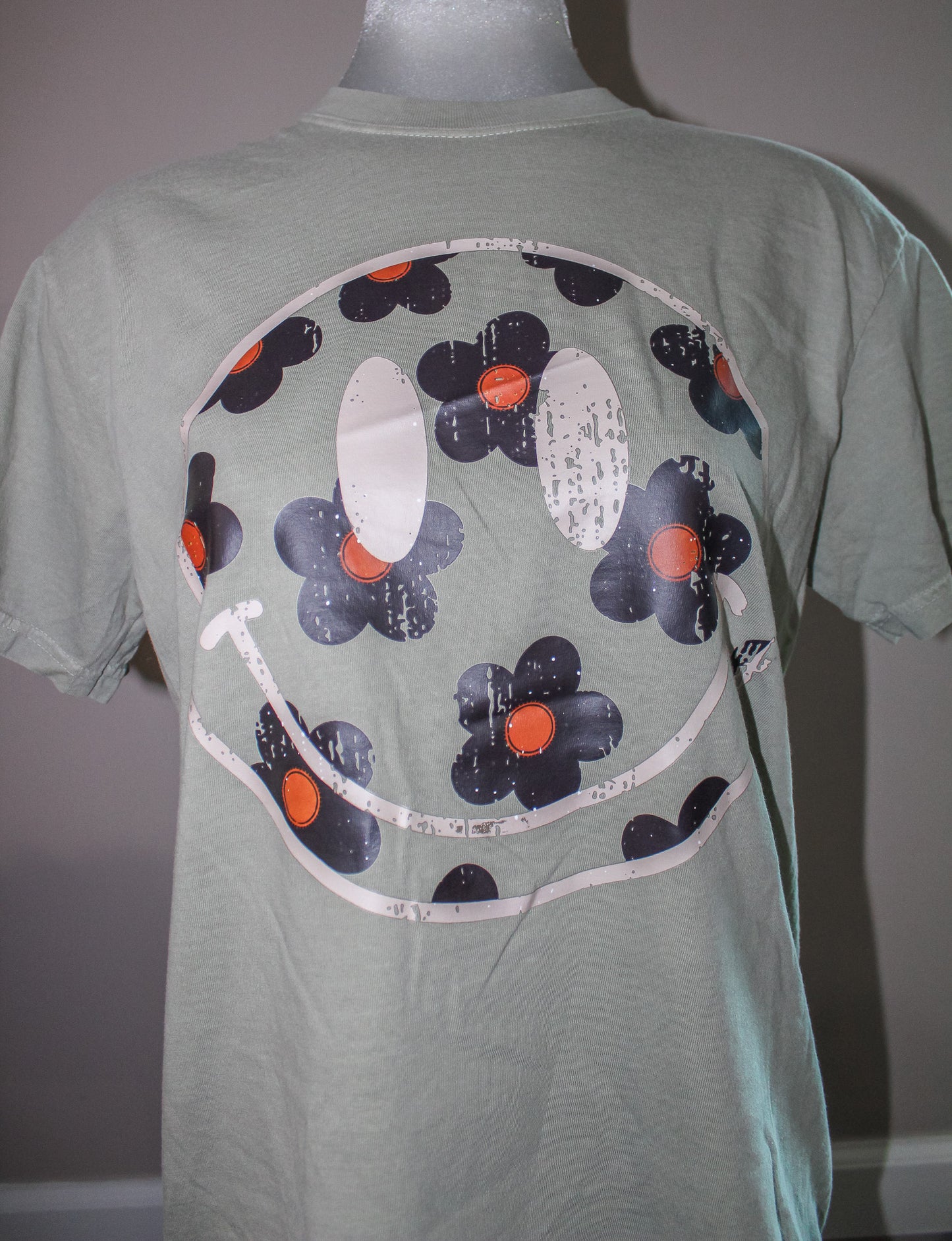 A sage green short-sleeve t-shirt displayed on a mannequin, featuring a large graphic print of a smiley face made of dark blue-gray flowers with orange centers and white distressed outlines.