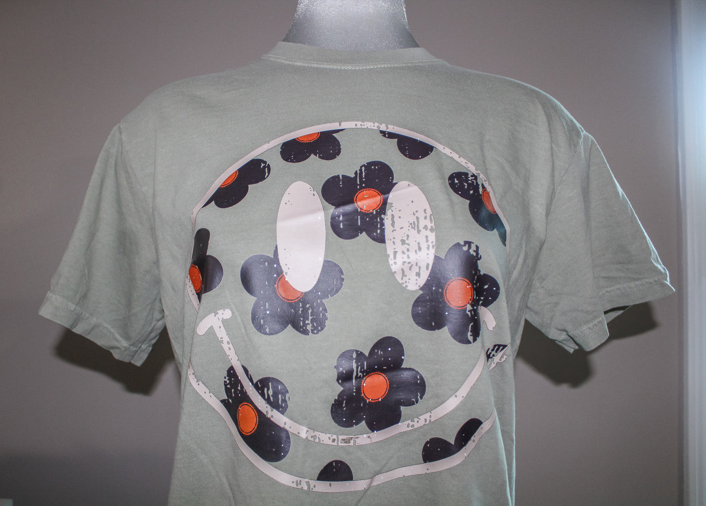 A sage green short-sleeve t-shirt displayed on a mannequin, featuring a large graphic print on the back with a smiley face made of dark blue-gray flowers with orange centers and white outlines.