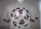A sage green short-sleeve t-shirt displayed on a mannequin, featuring a large graphic print on the back with a smiley face made of dark blue-gray flowers with orange centers and white outlines.