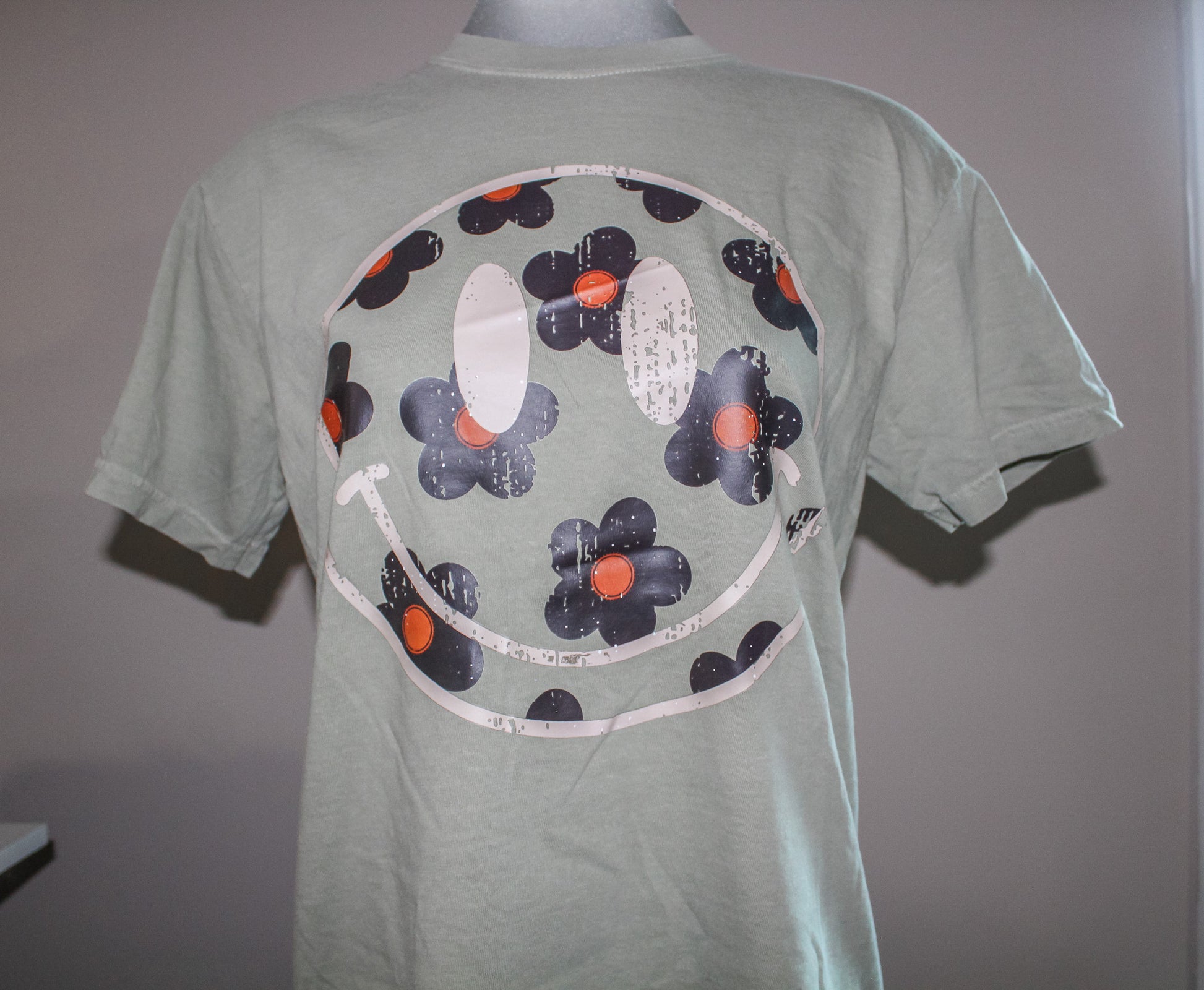 A light sage green short-sleeve t-shirt displayed on a mannequin torso, featuring a large circular graphic print on the back with stylized flowers in black, white, and orange.