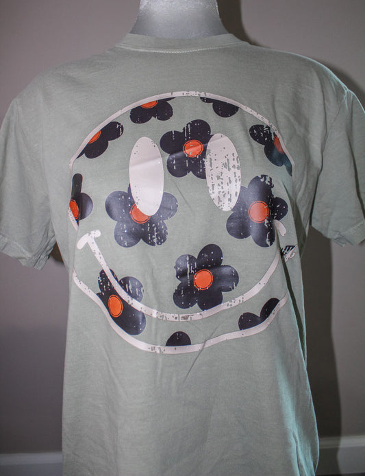 A sage green short-sleeve t-shirt displayed on a mannequin, featuring a large graphic print of a smiley face made of dark blue-gray flowers with orange centers and white distressed outlines.