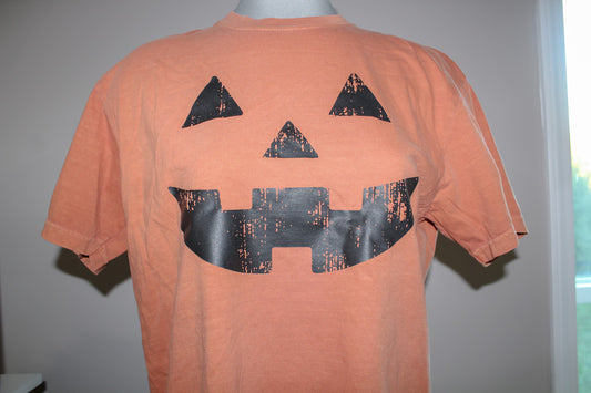 An orange short-sleeve t-shirt displayed on a mannequin torso, featuring a large black distressed jack-o'-lantern face graphic on the front.