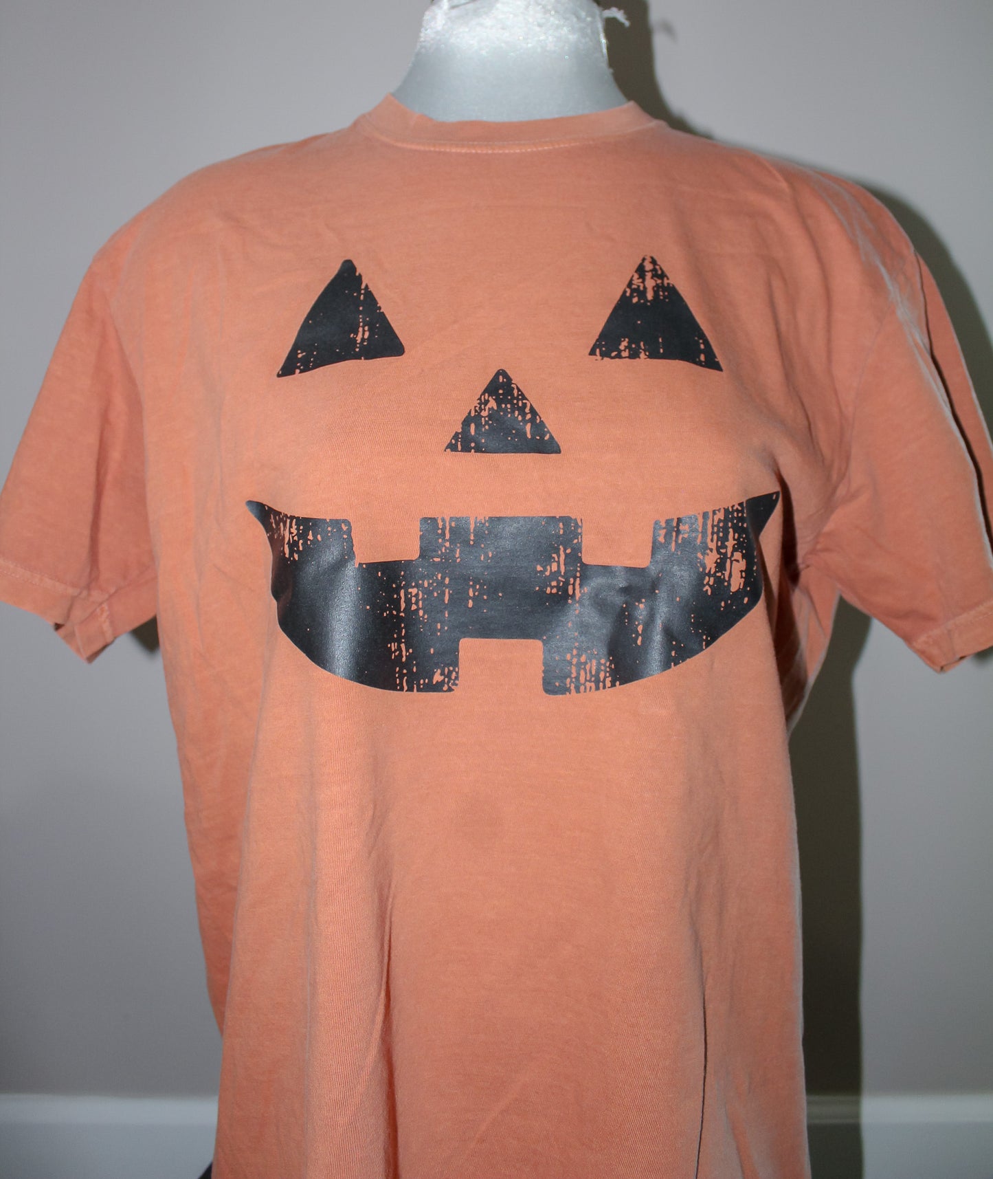 An orange short-sleeve t-shirt displayed on a mannequin torso, featuring a large black distressed jack-o'-lantern face graphic on the front.