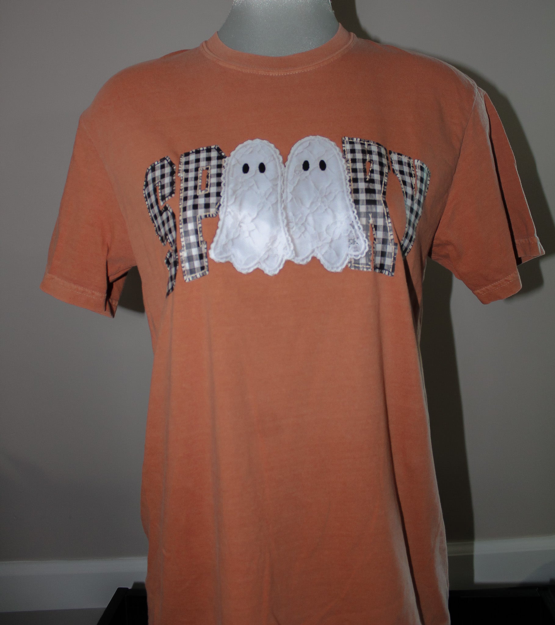 An orange short-sleeve t-shirt displayed on a mannequin torso, featuring a 'SPOIL' graphic across the chest with two white lace ghosts and black-and-white gingham patterned letters.