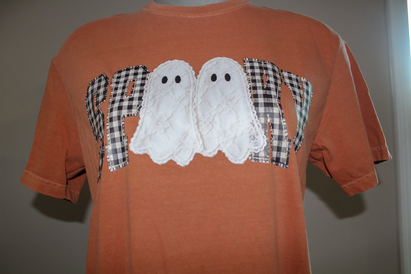 An orange short-sleeve t-shirt featuring a 'HAPPY' graphic across the chest, where the letters are filled with black-and-white gingham plaid and the center contains two white textured ghost appliqués with black eyes.