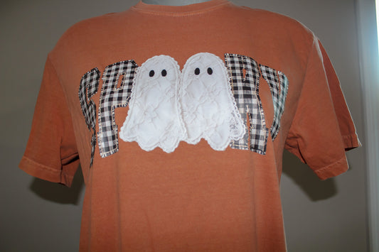 An orange short-sleeve t-shirt featuring a 'HAPPY' graphic across the chest, where the letters are filled with black-and-white gingham plaid and the center contains two white textured ghost appliqués with black eyes.