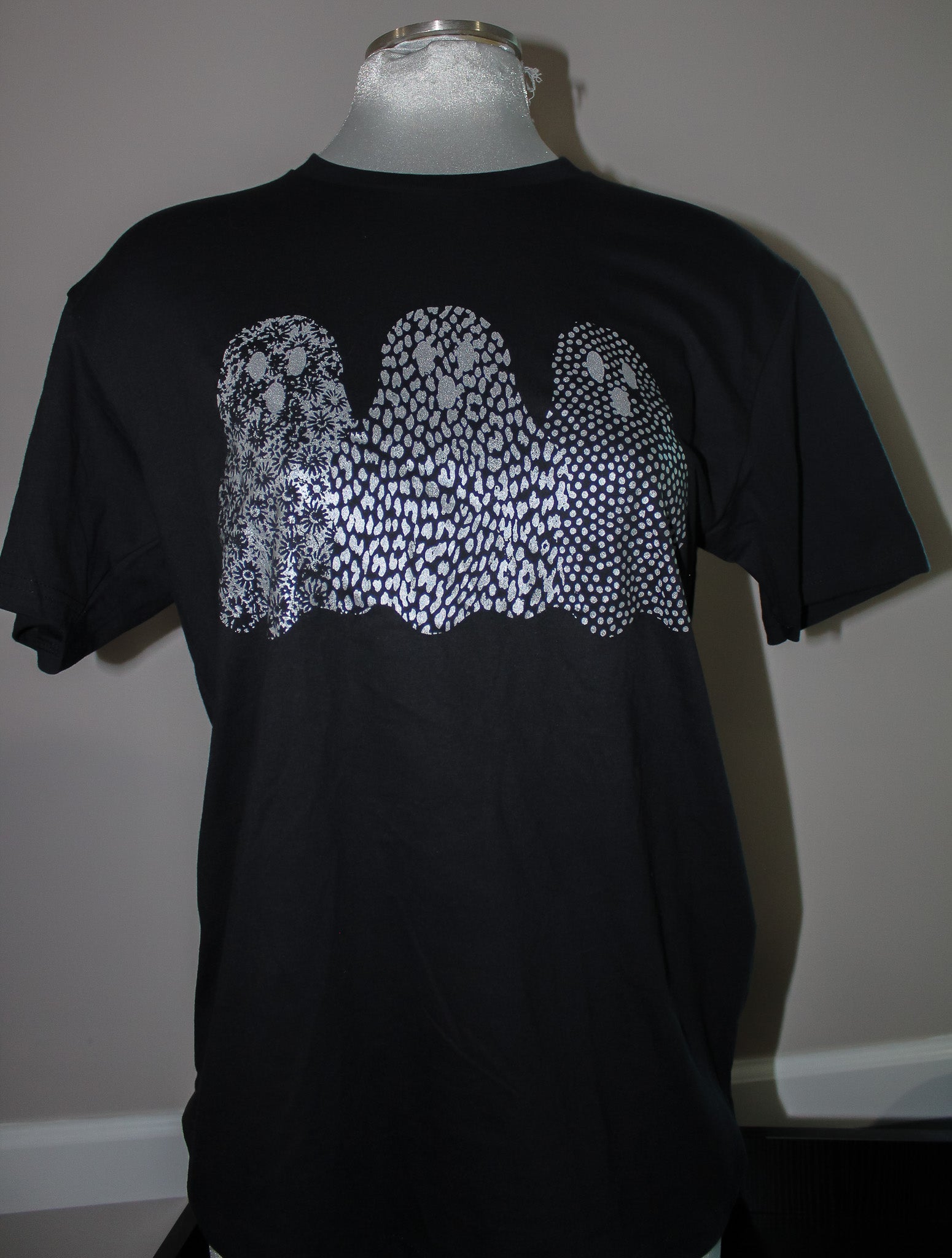 A black short-sleeve t-shirt displayed on a mannequin torso, featuring a large white graphic of three ghosts made of dense dots and lines across the chest.