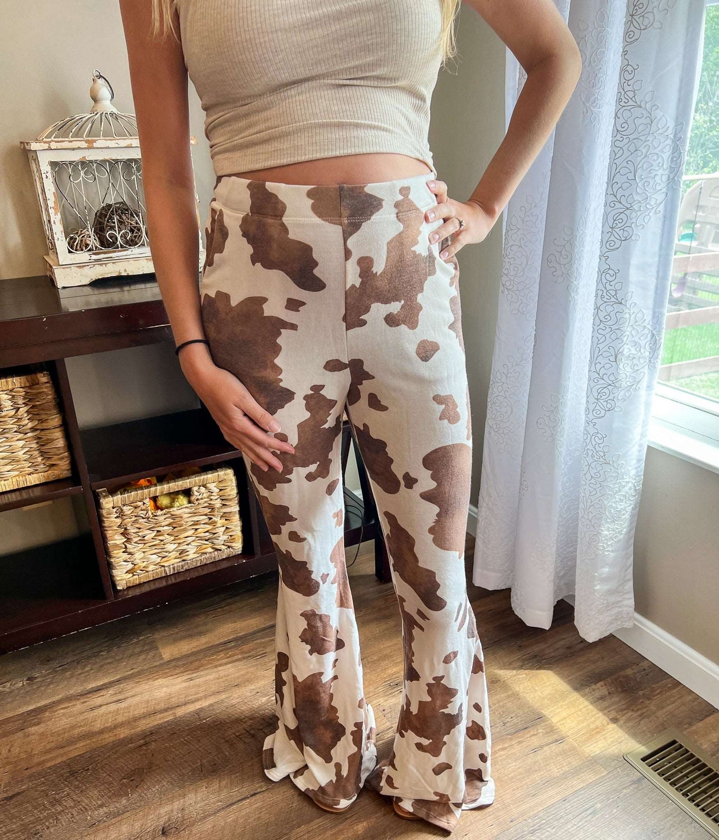 Jade by Jane High rise mocha cow print flare pants. Model wearing cow print flare pants and an oatmeal colored crop top for a trendy and casual outfit. Mocha Cow print pants available at Lexington and Gray sizes Small through 3x